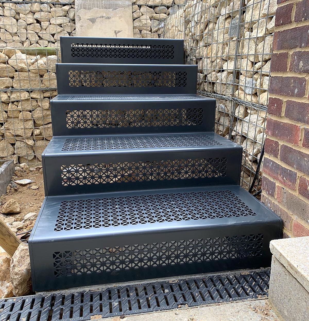 Perforated dark grey steel treads and risers climbing between two gabion walls