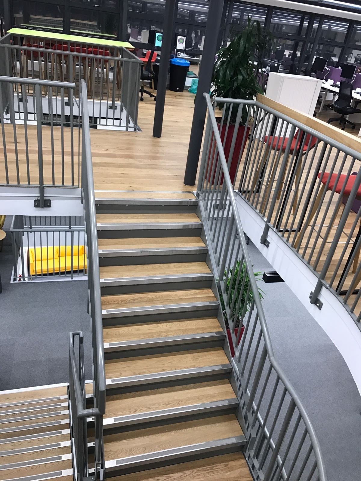 Office stair from the landing above: grey steel stringers carrying oak boards, upright bar sides